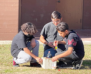 Three male students working on a bottle rocket on the school field